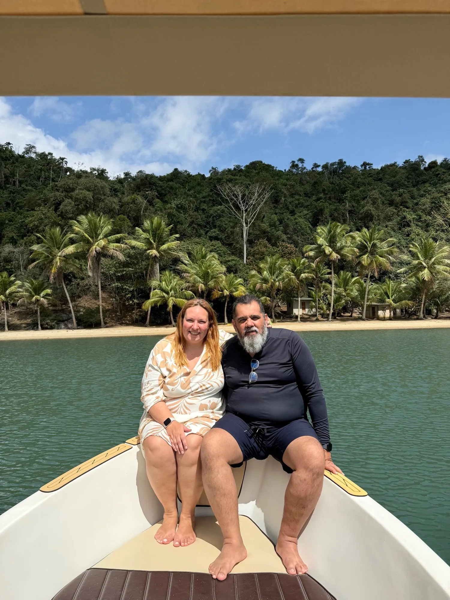Dérica e Luis sentados na proa de um barco, com uma praia de coqueiros ao fundo