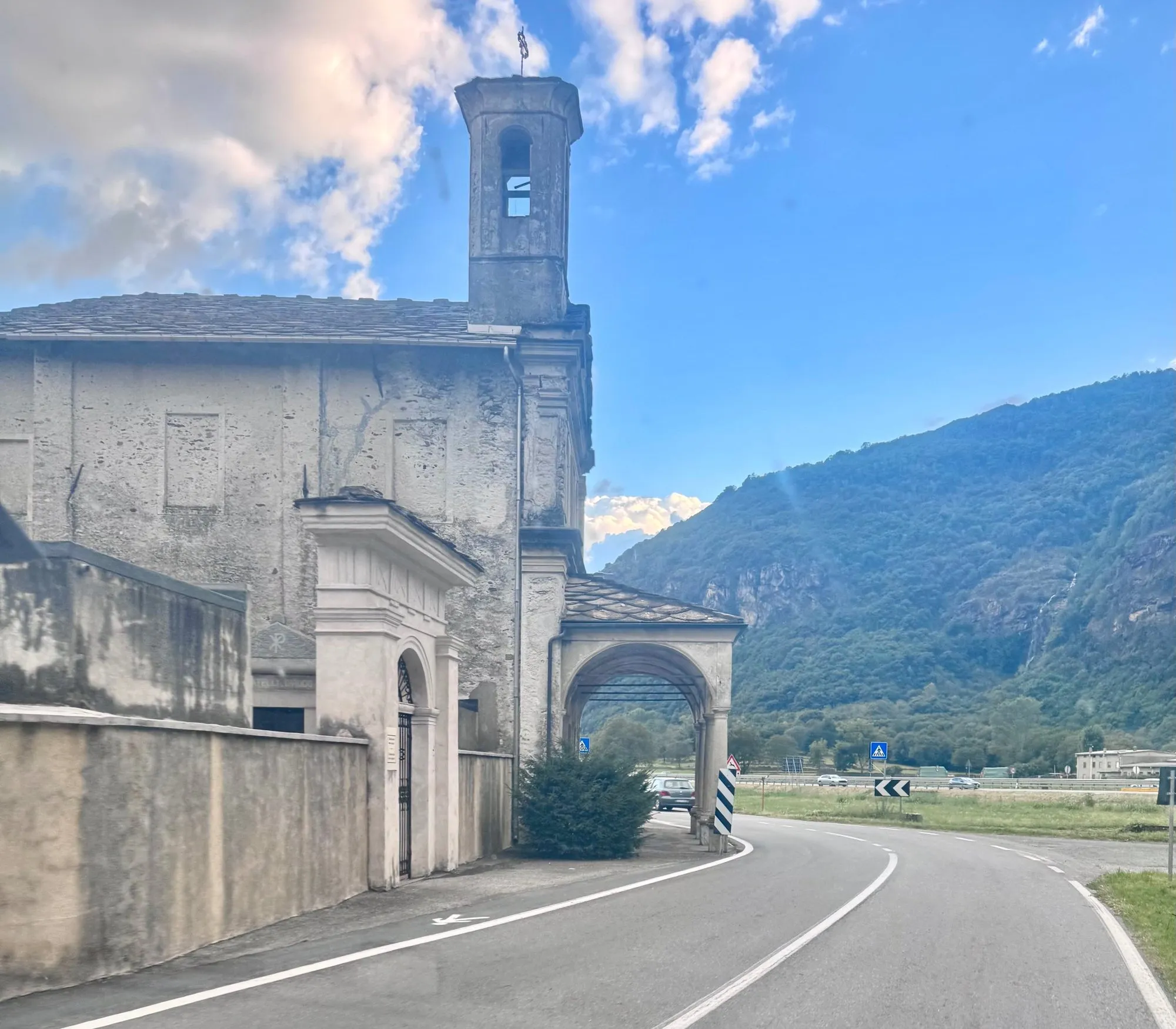 Igreja antiga à beira de uma estrada, com montanhas e céu azul ao fundo