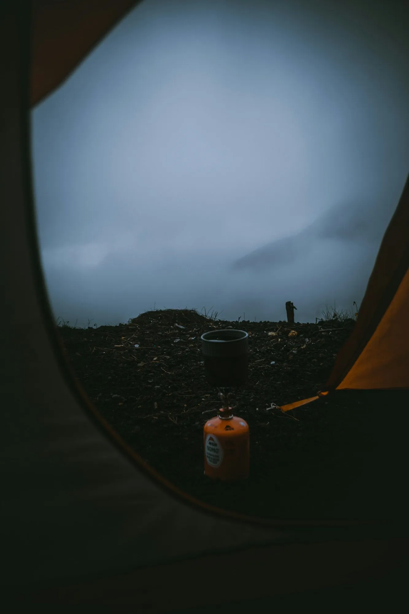Vista de dentro de uma barraca: fogareiro com caneca e céu nublado ao amanhecer